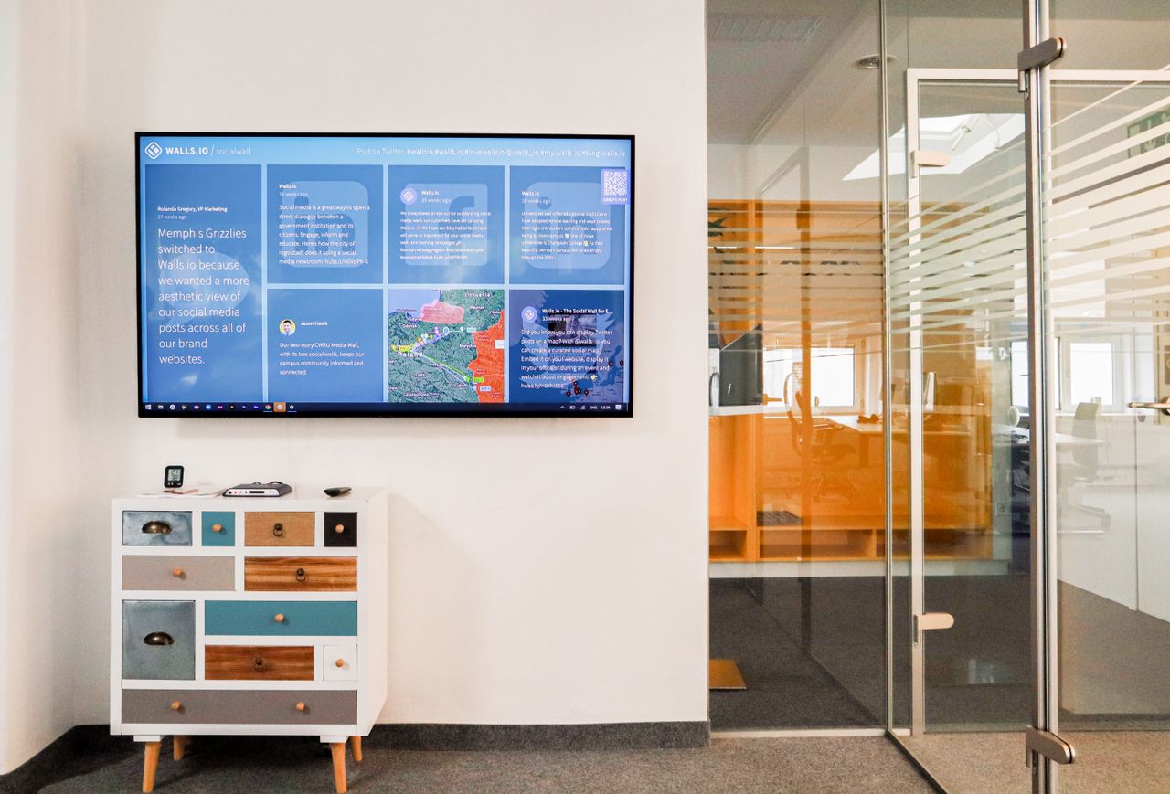 About Sleek office interior featuring a wall-mounted TV screen and colorful dresser in a glass-walled room.